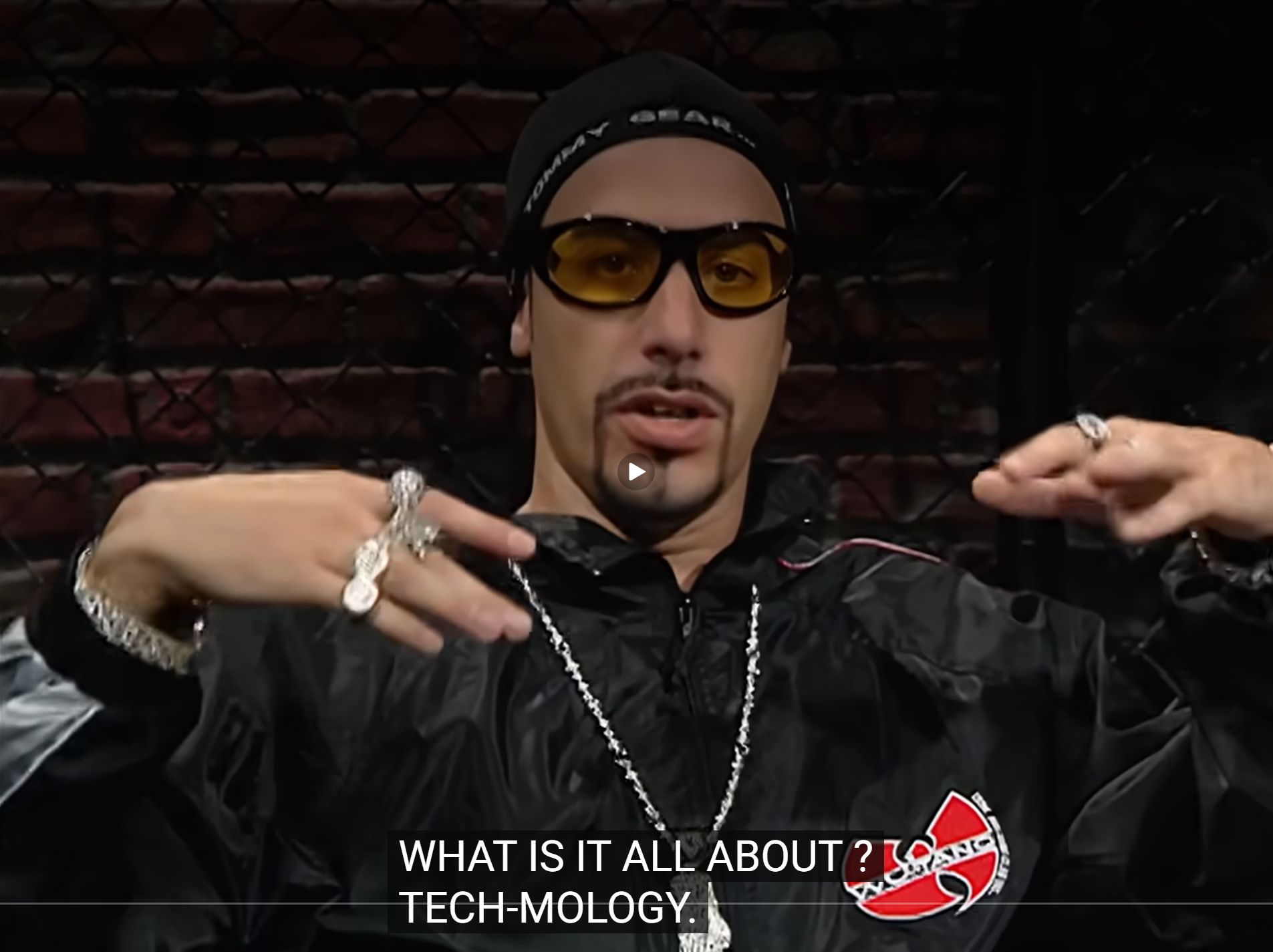 Sacha Baron Cohen in dark clothing and tinted glasses, captioned What is it all about? Tech-mology