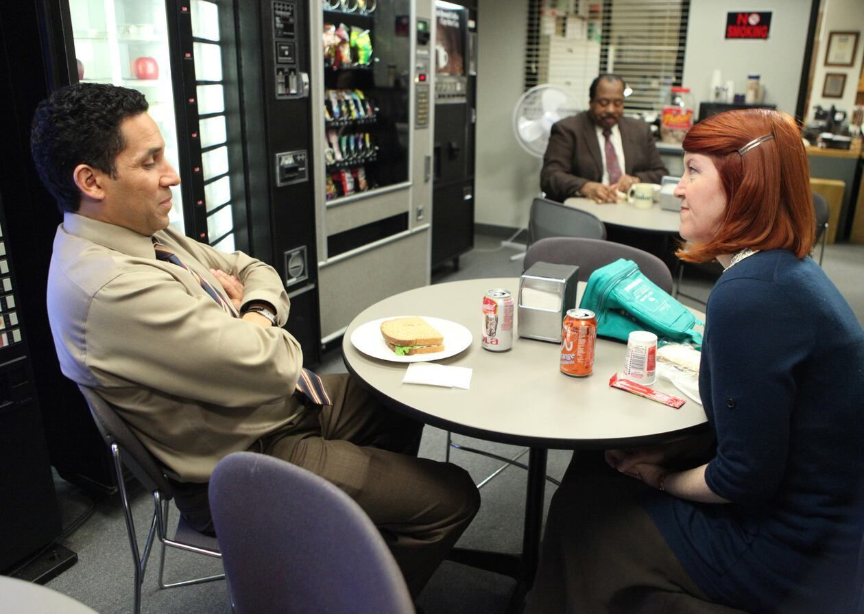 Coworkers talking across a small break-room table beside vending machines in a scene from The Office